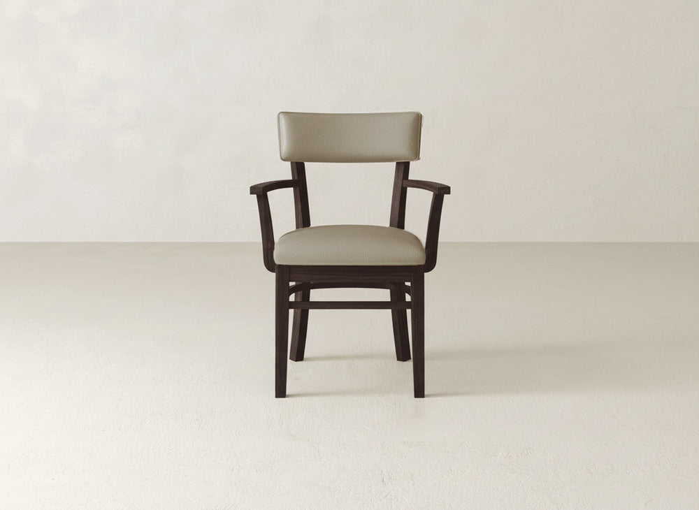 The Everly Arm Dining Chair by James+James, featuring a light brown cushioned seat and backrest with armrests and a simple mid-century modern design, is centered against a plain, light-colored wall and floor.