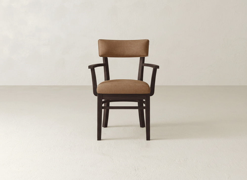 The Everly Arm Dining Chair by James+James features a brown cushioned seat and backrest with wooden armrests, set on a light-colored floor against an off-white wall.