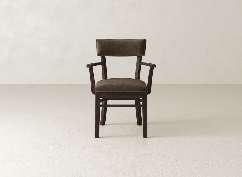 The Everly Arm Dining Chair by James+James, featuring a padded brown seat and backrest, is centered in an empty, light-colored room with a plain wall.