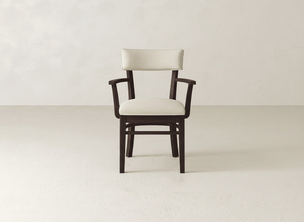 The Everly Arm Dining Chair by James+James features a mid-century modern design with armrests and a cushioned seat and backrest in light beige fabric, set against a plain, light-colored wall and floor.