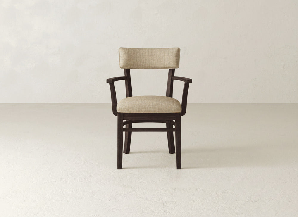 The Everly Arm Dining Chair by James+James features a wooden frame and light beige upholstered seat and backrest with armrests, showcased against a plain, light-colored background in a mid-century modern style.