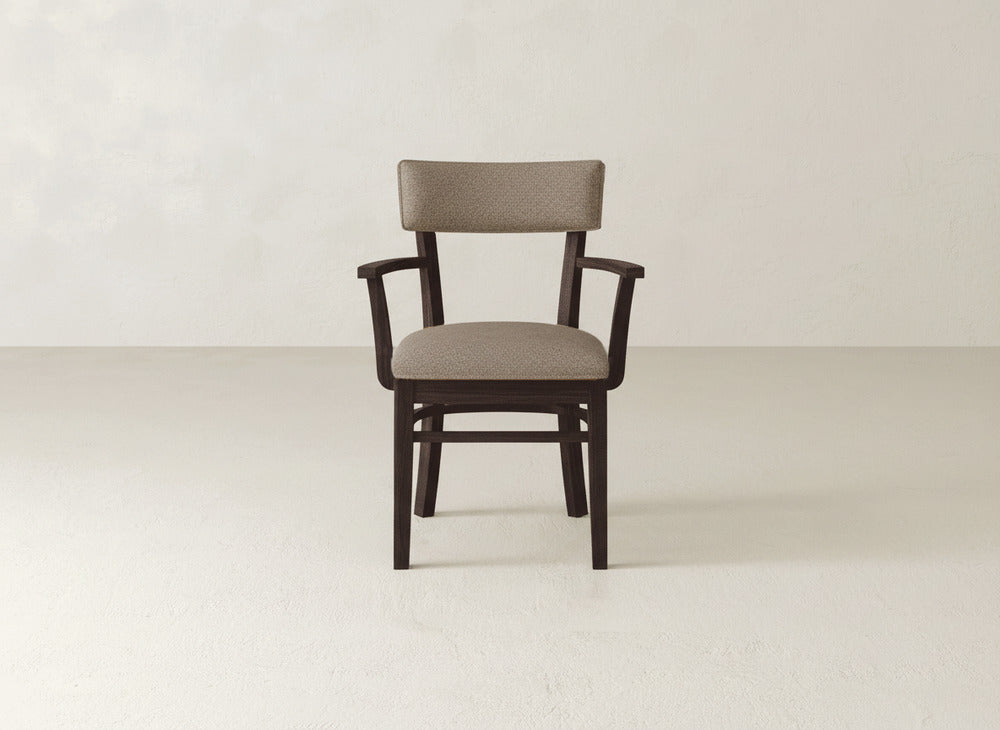 The Everly Arm Dining Chair by James+James features a brown upholstered seat and backrest with wooden armrests, elegantly set on a light-colored floor against an off-white wall.