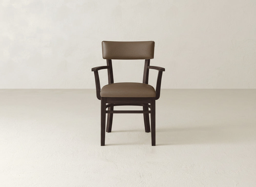 The Everly Arm Dining Chair by James+James, featuring a wooden frame, armrests, and brown cushioned seat and backrest, sits on a light floor against a plain beige wall.