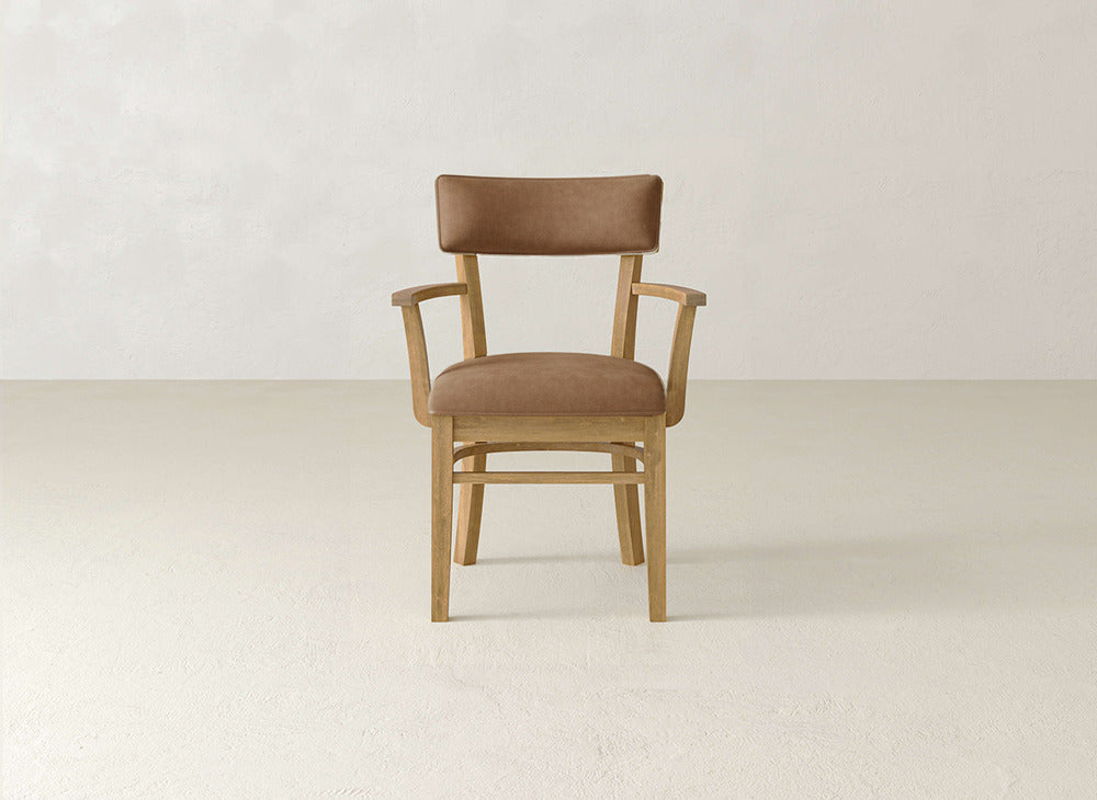 The Everly Arm Dining Chair by James+James, featuring a light brown upholstered seat and back with wooden armrests, is placed on a light floor against an off-white wall.