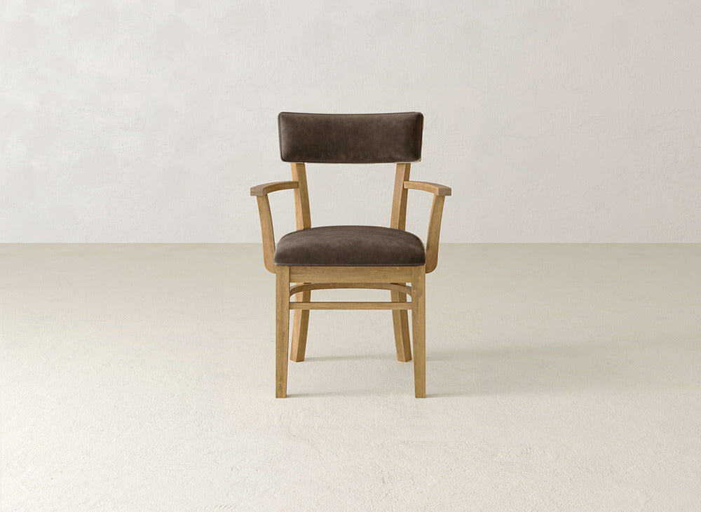 The Everly Arm Dining Chair by James+James, featuring brown upholstery and wooden armrests, is placed on a light floor against an off-white wall.