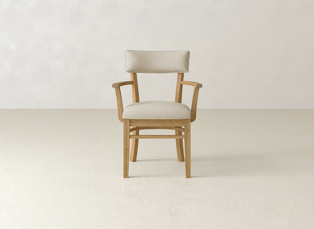 The Everly Arm Dining Chair by James+James features a light beige upholstered seat and backrest, simple minimalist design, and comfortable armrests. It sits on a plain floor against a textured off-white wall, highlighting its mid-century modern style.