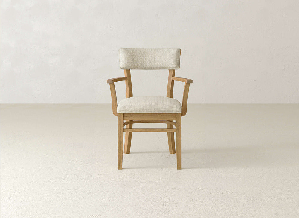 The Everly Arm Dining Chair by James+James, featuring a light beige upholstered seat, backrest, and padded armrests, is placed in a minimal, light-toned room with textured walls and floors.