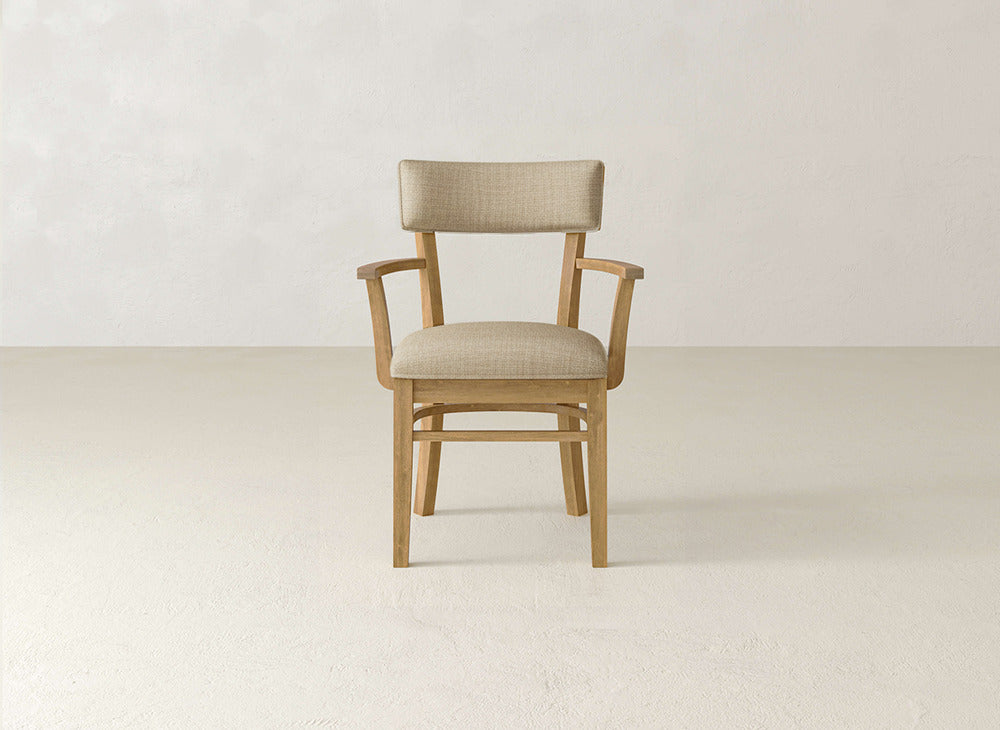 The Everly Arm Dining Chair by James+James, featuring a light beige upholstered seat and backrest with wooden armrests, is placed against a plain, light-colored wall and floor.