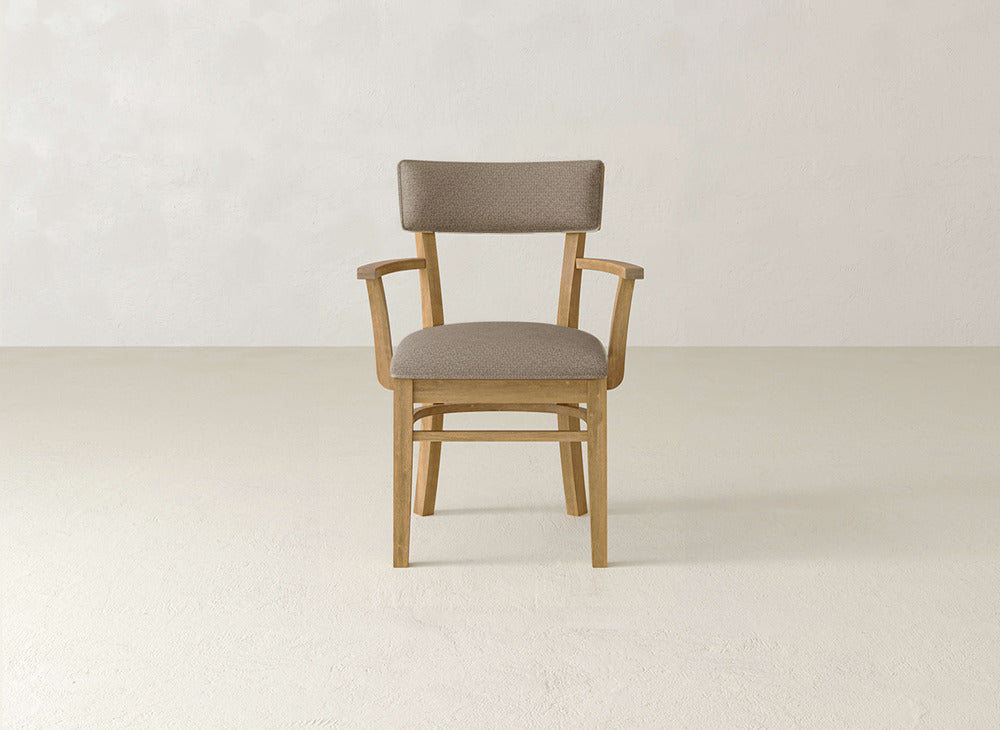The Everly Arm Dining Chair by James+James features a wooden frame, light brown cushioned seat and backrest, simple armrests, and is placed on a light-colored floor against an off-white wall.