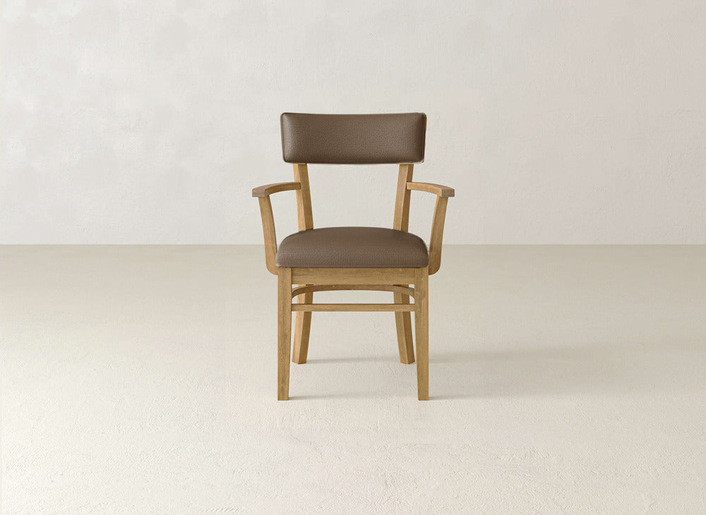The Everly Arm Dining Chair by James+James features a wooden frame and brown cushioned seat and backrest, positioned on a light floor against a plain, light wall.