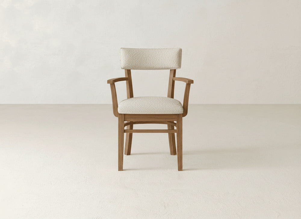 The Everly Arm Dining Chair by James+James, featuring a light beige upholstered seat and backrest, is placed on a light floor against a plain off-white wall.