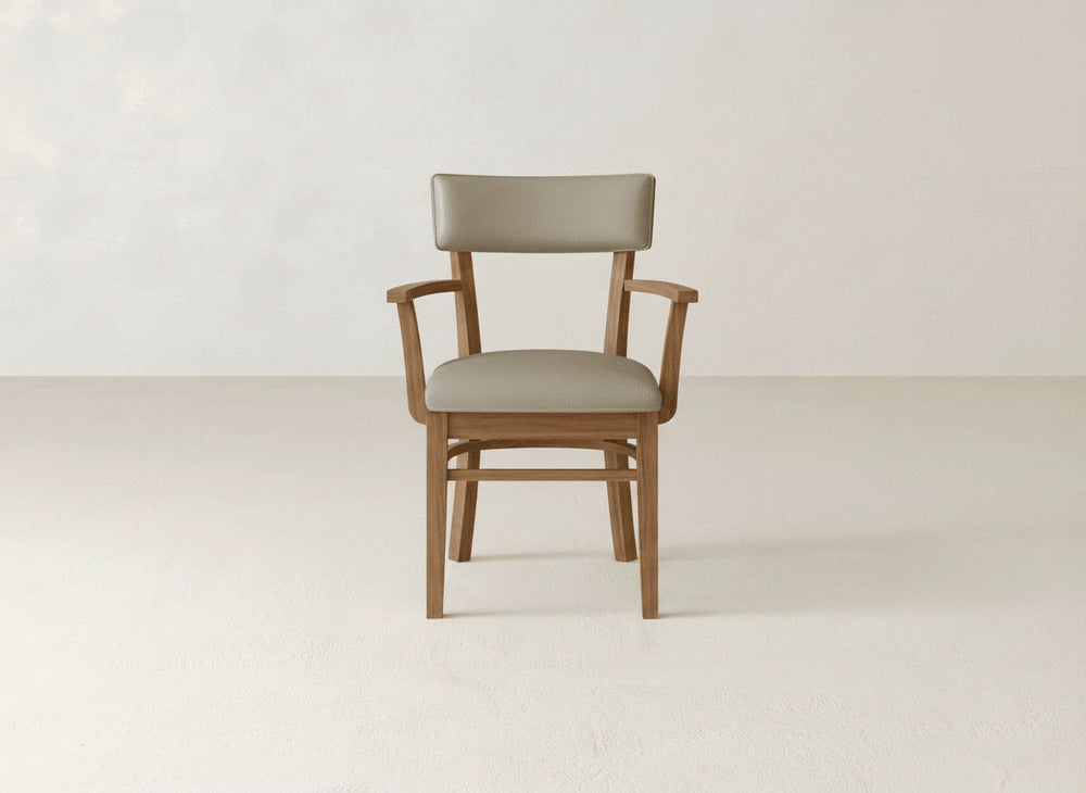 The Everly Arm Dining Chair by James+James features armrests and a light brown upholstered seat and backrest, adding mid-century modern elegance to a minimal, light-colored room with plain walls and floor.