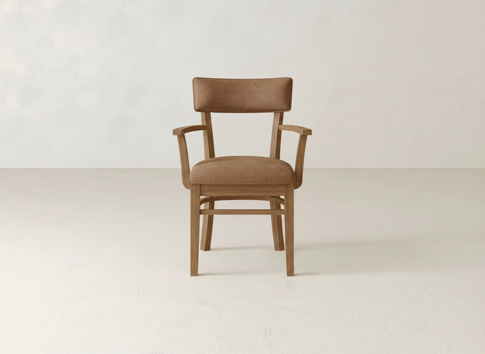 The Everly Arm Dining Chair by James+James features light brown upholstered seat and backrest with armrests, shown against a plain light-colored wall and floor.