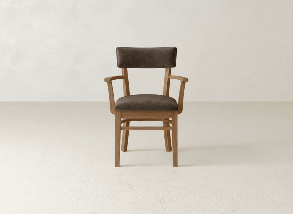 The Everly Arm Dining Chair by James+James features a dark brown upholstered seat and backrest with armrests, displayed on a light floor against a neutral wall.