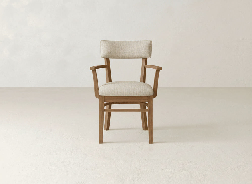 The Everly Arm Dining Chair by James+James features a cushioned beige seat and backrest with armrests, displayed on a light floor against a neutral, textured wall.