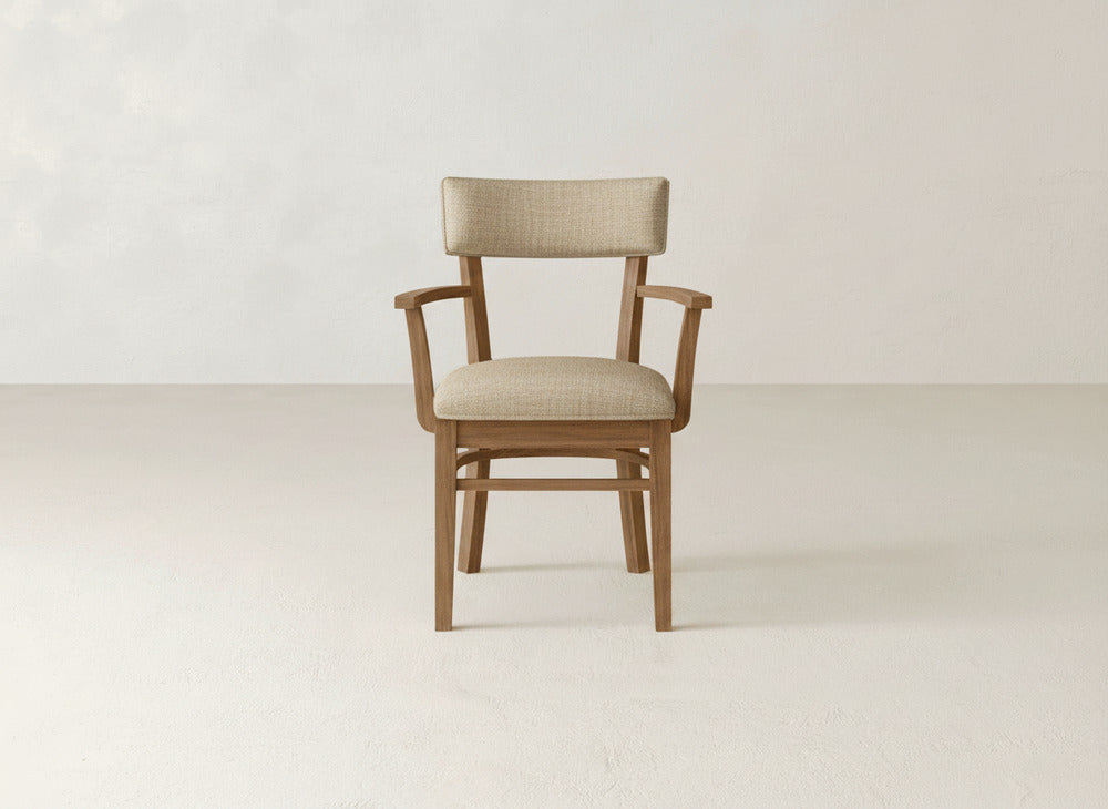 The Everly Arm Dining Chair by James+James, featuring a beige upholstered backrest and seat, is showcased in a minimalist room with light tones, smooth floors, and plain walls.