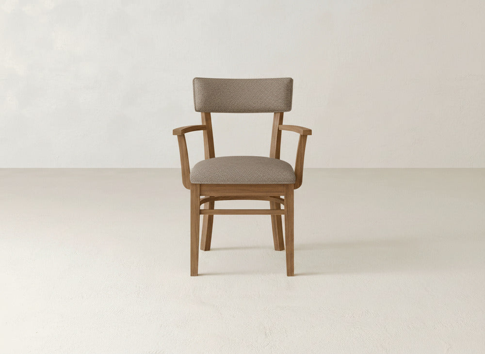 The James+James Everly Arm Dining Chair, with armrests and a padded brown seat and backrest, sits in an empty room with a light floor and plain off-white wall.