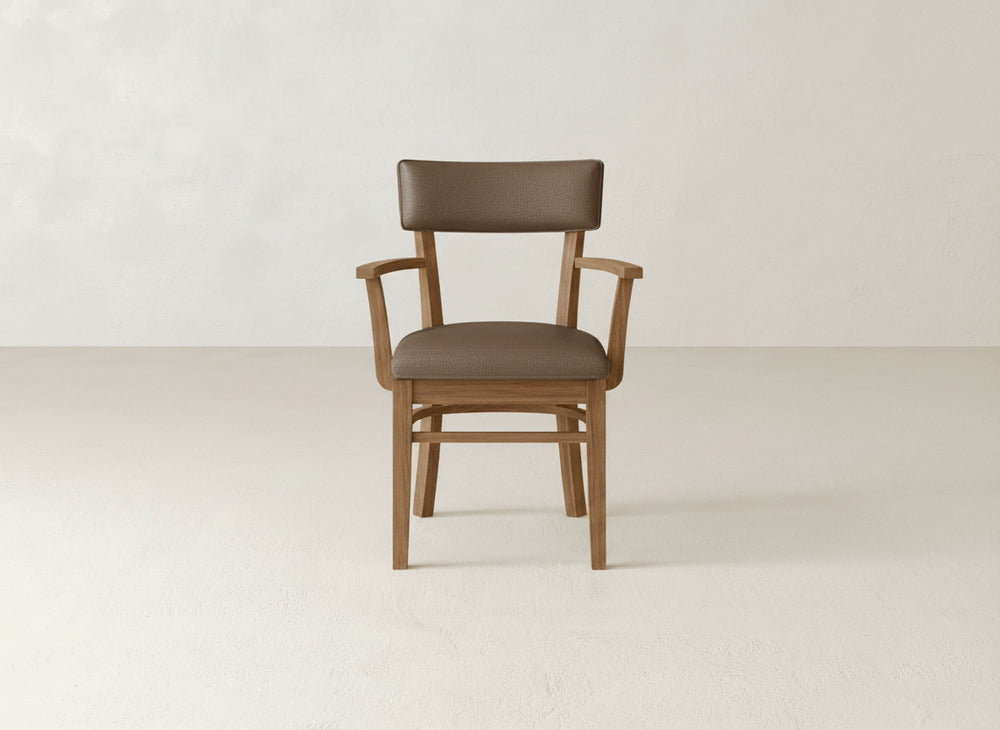 The Everly Arm Dining Chair by James+James, a mid-century modern piece with a brown cushioned seat and wooden armrests, is set against a plain light-colored wall and floor.