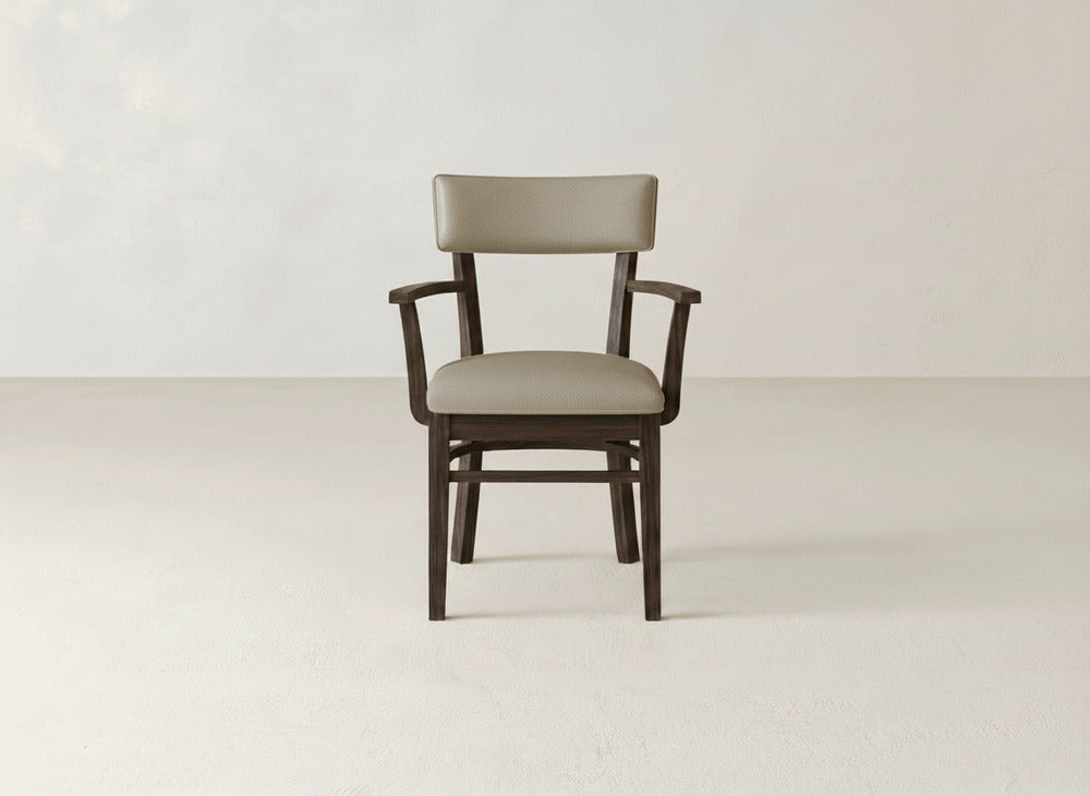 The Everly Arm Dining Chair by James+James, with taupe upholstery and dark wood frame and legs, stands centered against a plain light wall and floor.