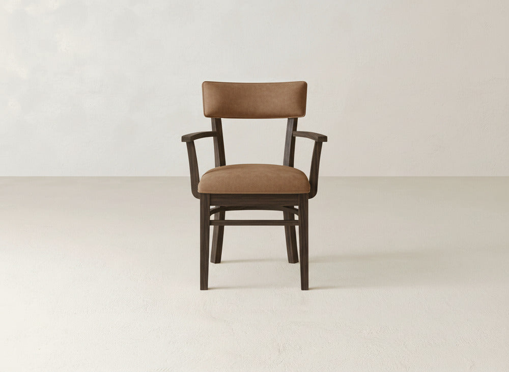 The James+James Everly Arm Dining Chair, featuring brown upholstery and a wooden frame, sits on a light floor against a plain off-white wall.