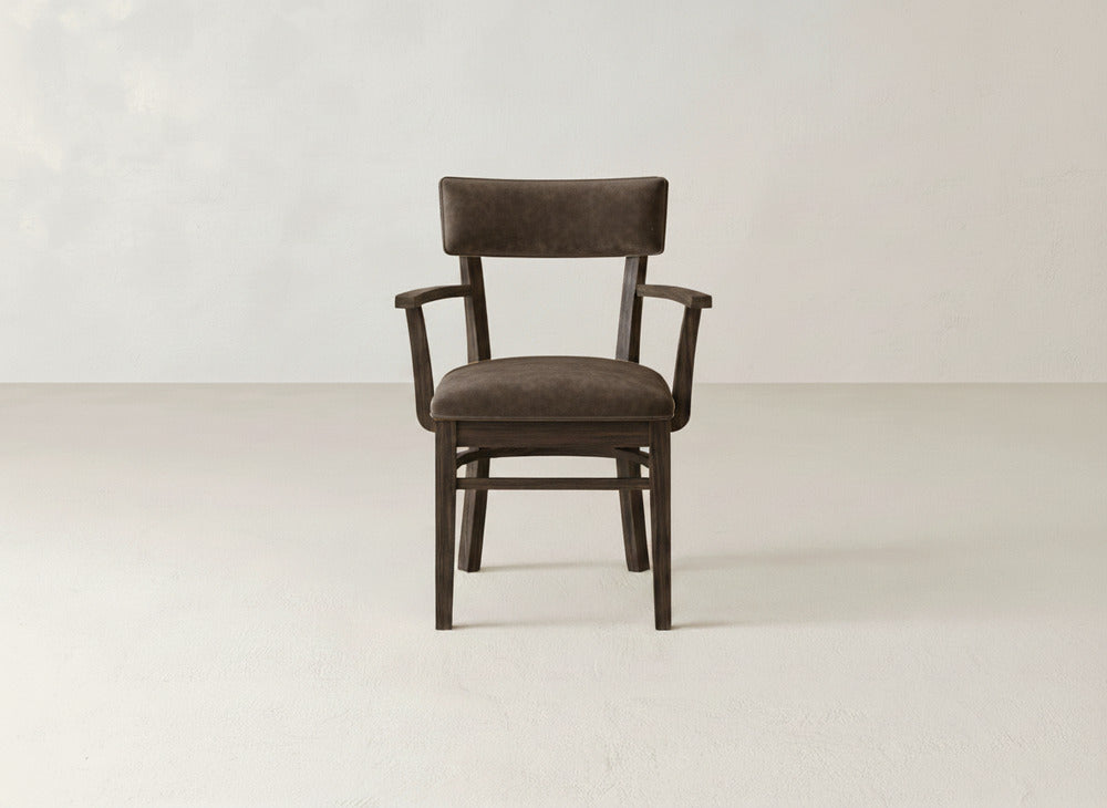 The Everly Arm Dining Chair by James+James, a dark brown mid-century modern chair with padded seat, backrest, and armrests, is centered facing forward on a light floor against a plain light background.