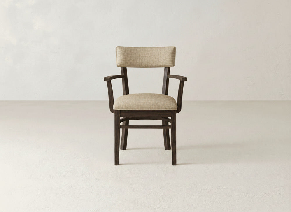The Everly Arm Dining Chair by James+James, featuring elegant upholstery with beige seat and backrest, is centered in an empty room with a light floor and plain wall background.