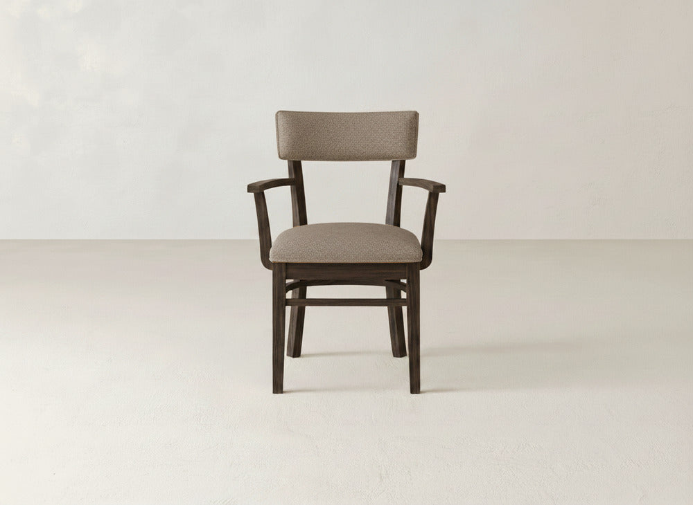 The Everly Arm Dining Chair by James+James, featuring a light brown upholstered seat and backrest with straight armrests and four legs, is set in a minimal, light-toned room with plain walls and flooring.