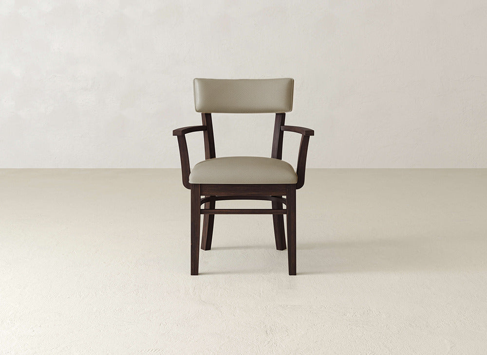 The James+James Everly Arm Dining Chair, featuring armrests and a light brown cushioned seat and backrest, is positioned on a smooth, light-colored floor against an off-white wall.