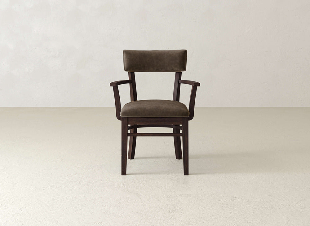 The Everly Arm Dining Chair by James+James, featuring dark wood and brown upholstered seat and backrest, sits in the center of a simple, light-colored room with bare walls and floor.