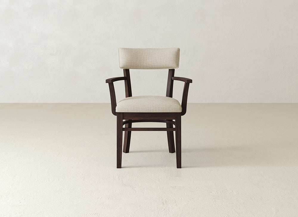 The Everly Arm Dining Chair by James+James features a cushioned seat and backrest in light beige fabric, armrests, and a simple modern design. Shown against a plain light background.