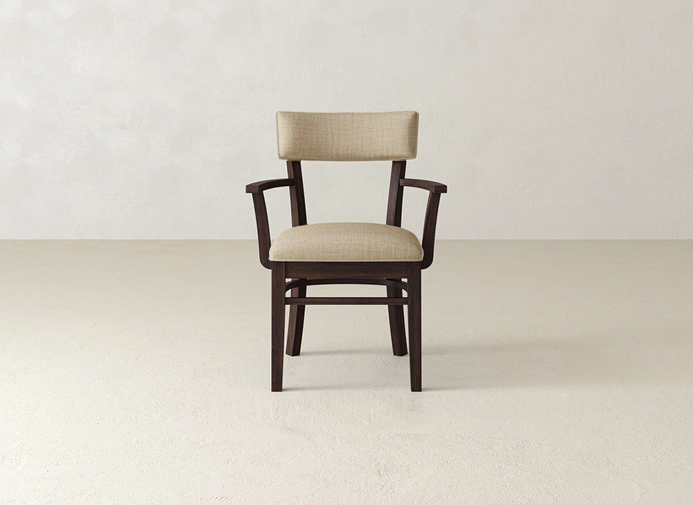 The Everly Arm Dining Chair by James+James, a mid-century modern piece with cushioned beige seat and backrest plus armrests, is centered in an empty room with light walls and floor.