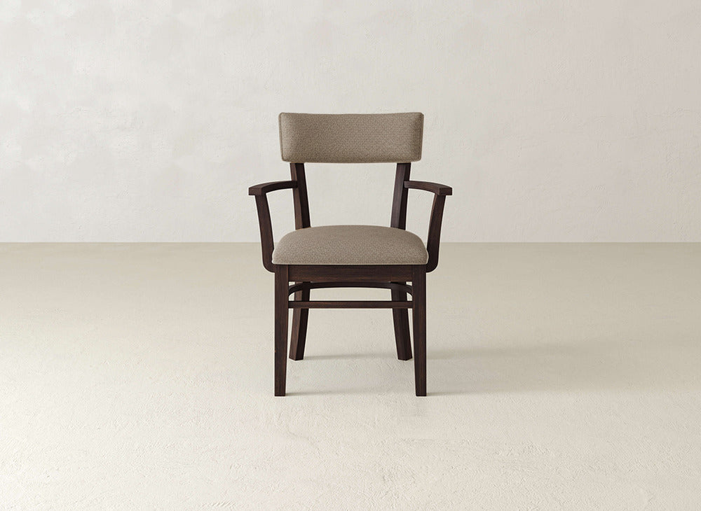 The Everly Arm Dining Chair by James+James, featuring a beige upholstered seat and backrest with armrests, is centered in a neutral-toned room with a plain wall and floor.