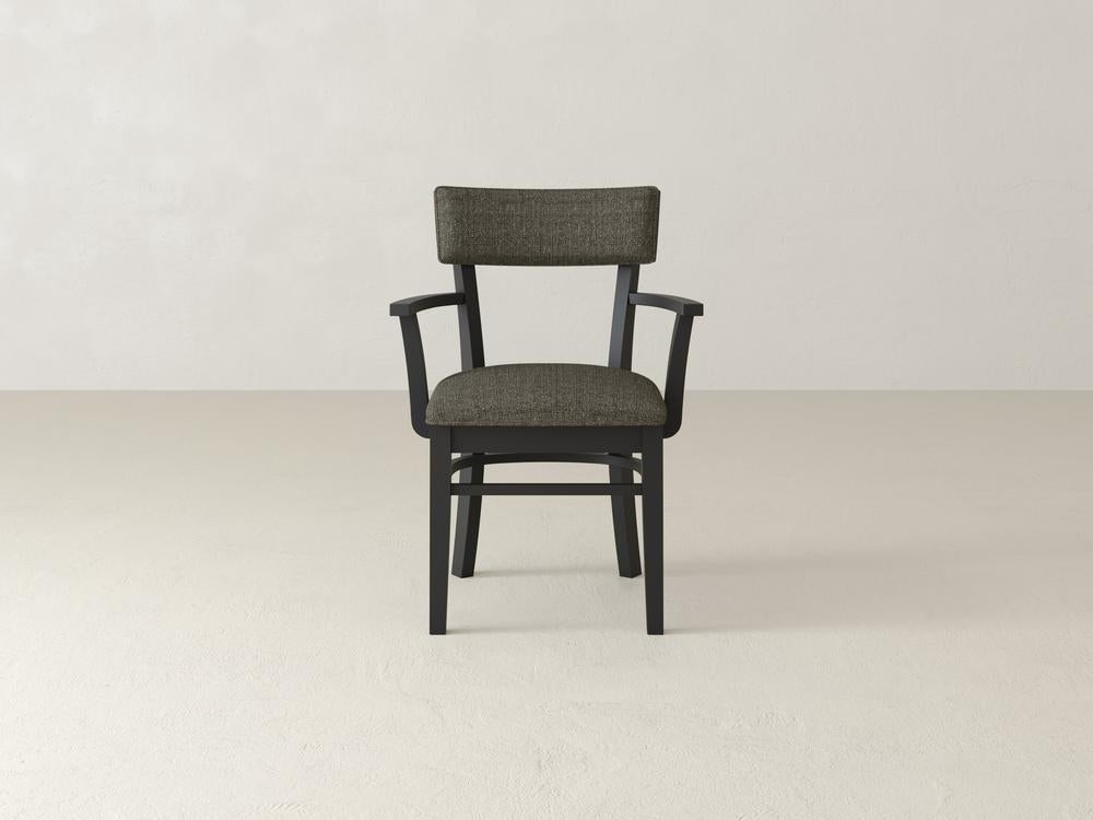 A wooden chair with a dark cushion on the seat and backrest is placed on a plain, light-colored floor against a neutral wall. The chair features simple armrests and a classic design.
