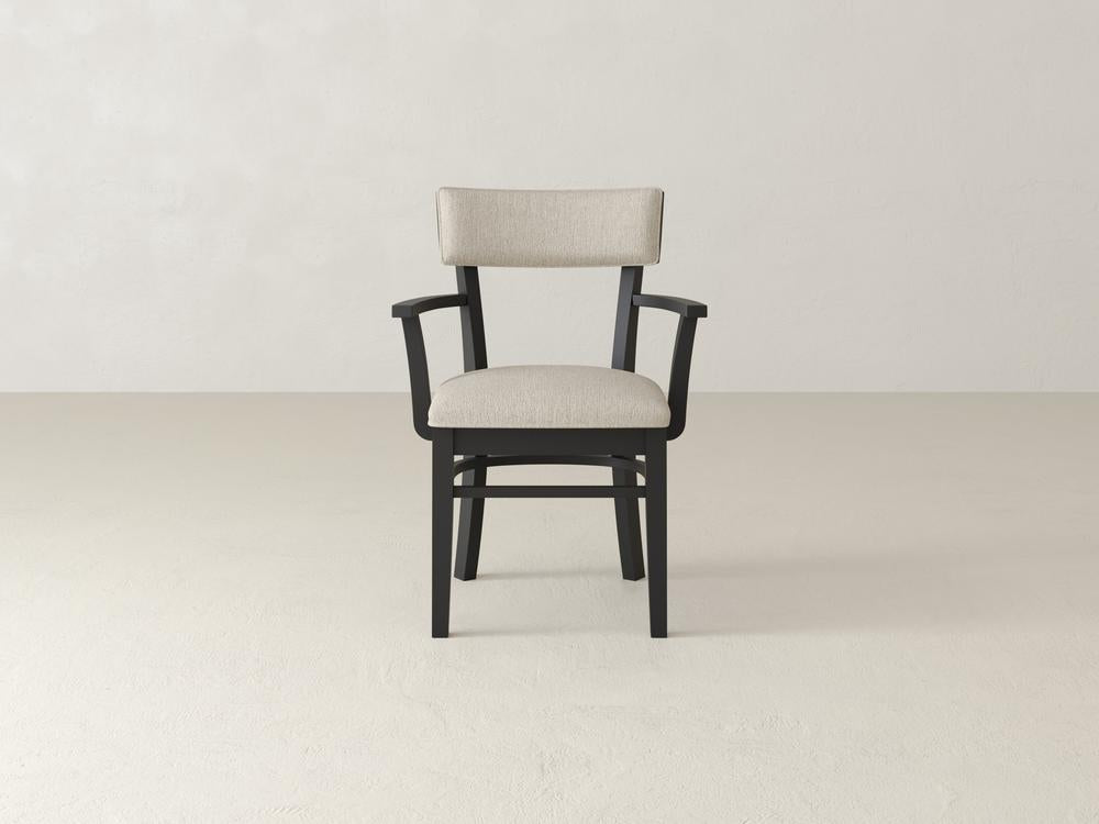A single wooden chair with a black frame and light beige padded seat and backrest is centered against a minimalist, light-colored background. The chair has armrests and a simple, modern design.