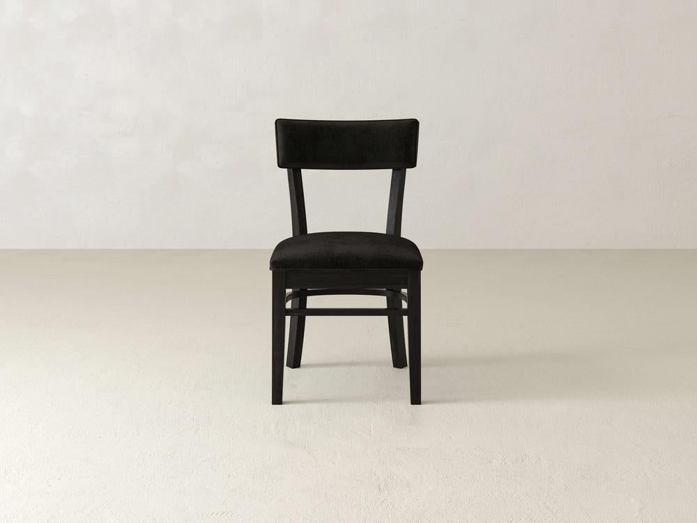 A black wooden chair with a cushioned backrest and seat is placed in the center of a plain, light-colored room. The floor and walls are a soft beige color, creating a minimalist and clean aesthetic.