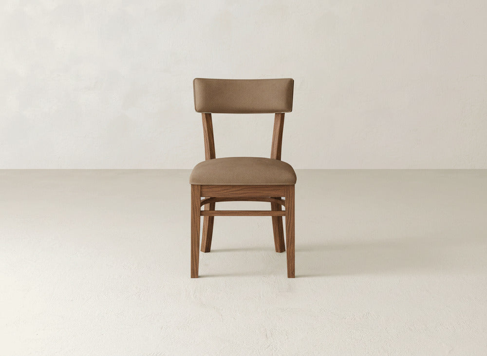 The Everly Side Dining Chair by James+James, featuring a brown upholstered seat and backrest, is centered in a minimalist room with light walls and floors.