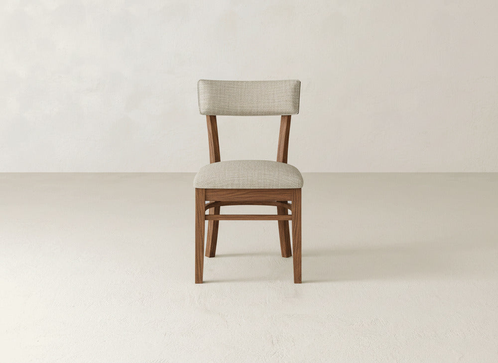 The James+James Everly Side Dining Chair, featuring a light beige cushioned seat and backrest, is centered in a minimalist room with a light-colored floor and plain textured wall.