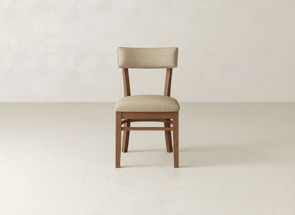 The Everly Side Dining Chair by James+James, featuring a light beige upholstered seat and backrest, is centered against a plain, light-toned wall and floor background.