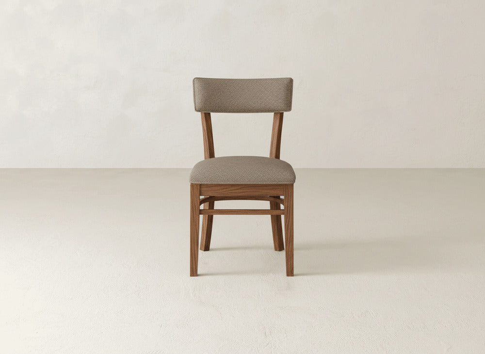The Everly Side Dining Chair by James+James features a wooden frame and cushioned seat and backrest in light brown fabric, standing alone on a light floor against a plain, off-white wall.
