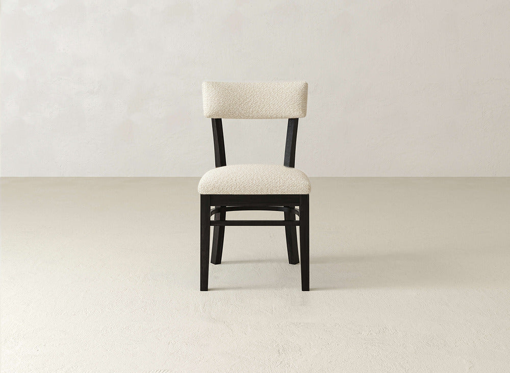 The Everly Side Dining Chair by James+James features a light beige cushioned seat and backrest with a dark wooden frame, and is centered against a plain light-colored wall and floor.