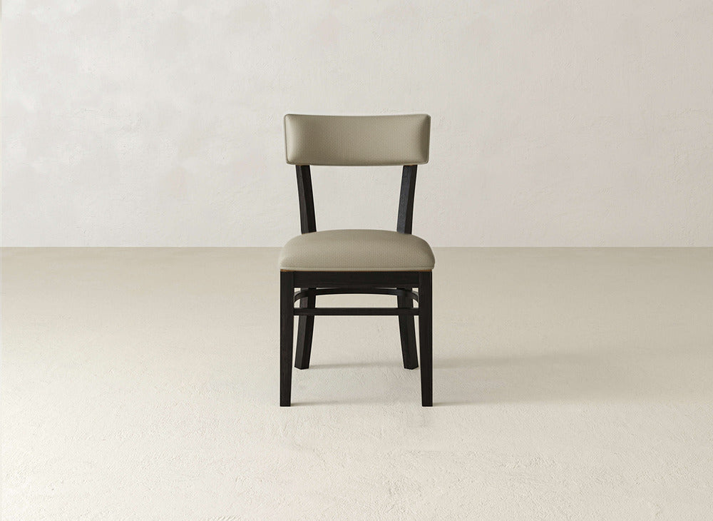 The Everly Side Dining Chair by James+James features a simple modern design with a light beige padded seat and backrest. Shown facing forward, it’s photographed against a plain, light-colored wall and floor.