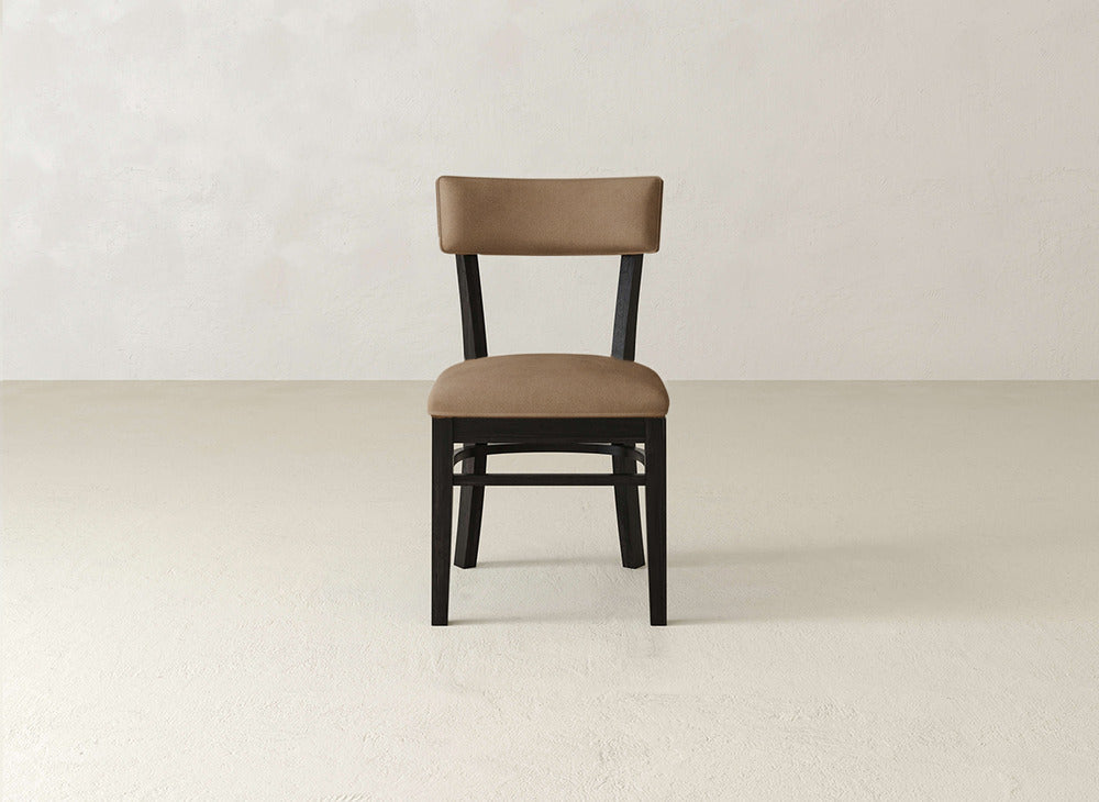 The Everly Side Dining Chair by James+James, featuring brown upholstery, a straight backrest, and black wooden legs, is centered against a plain, light-colored wall and floor.