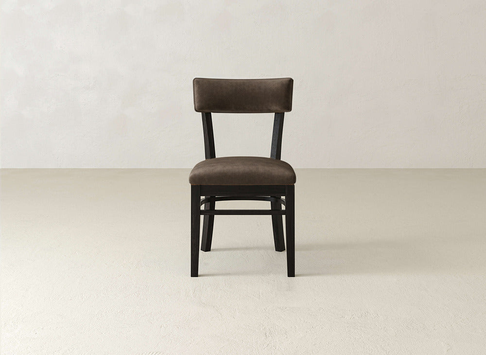 The Everly Side Dining Chair by James+James, with its brown upholstery, straight backrest, and dark wooden legs, is centered against a light wall and floor for a minimalist look ideal for modern dining spaces.