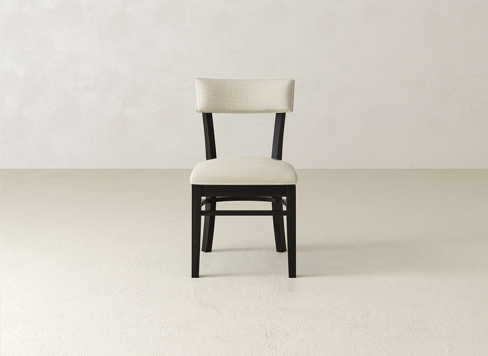 The Everly Side Dining Chair by James+James features a dark frame and light beige cushioned seat and backrest, creating a simple, modern, and minimalist look against a plain, light-colored wall and floor.