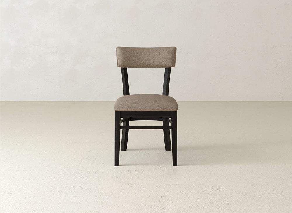 The Everly Side Dining Chair by James+James, featuring a cushioned seat and backrest in light brown fabric, is centered against a plain, light-colored wall and floor.