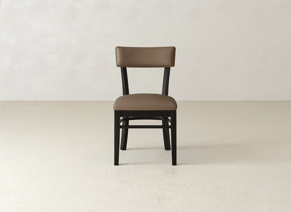 The Everly Side Dining Chair by James+James, featuring brown upholstery and a black wooden frame, is centered in a simple, light-toned room with an uncluttered background.
