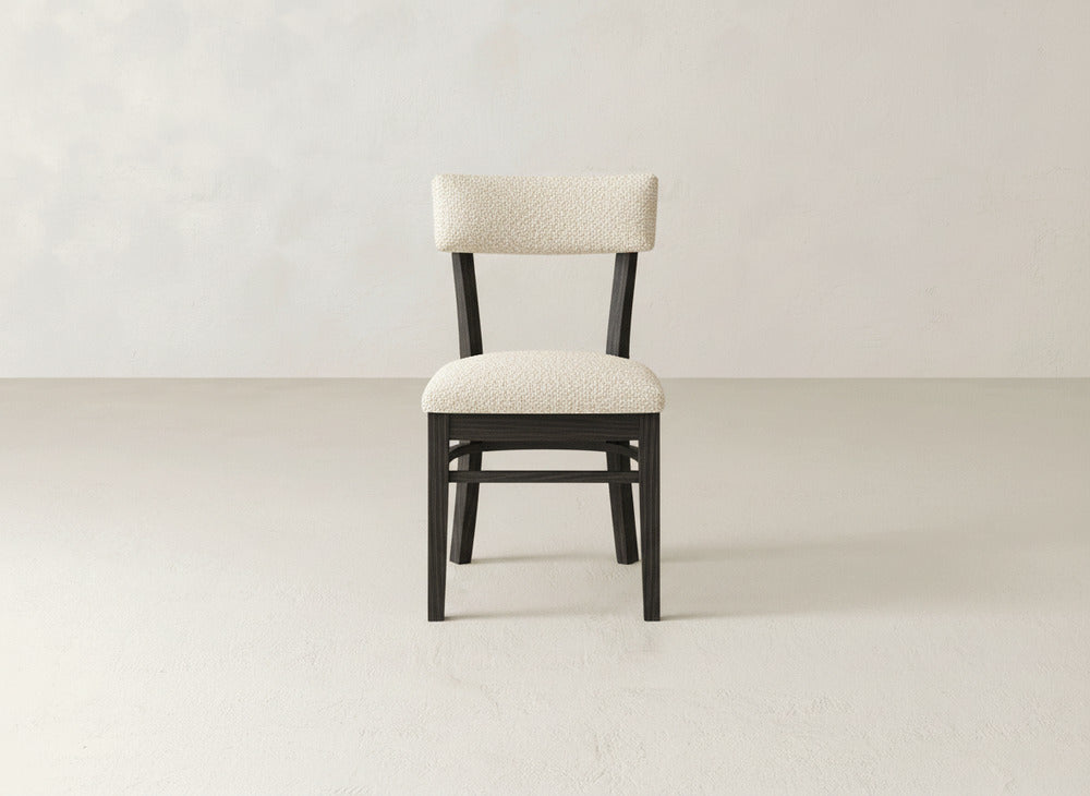 The Everly Side Dining Chair by James+James features a black frame and light, textured upholstered seat and backrest. It sits centered on a plain, light-colored floor with a neutral wall background.