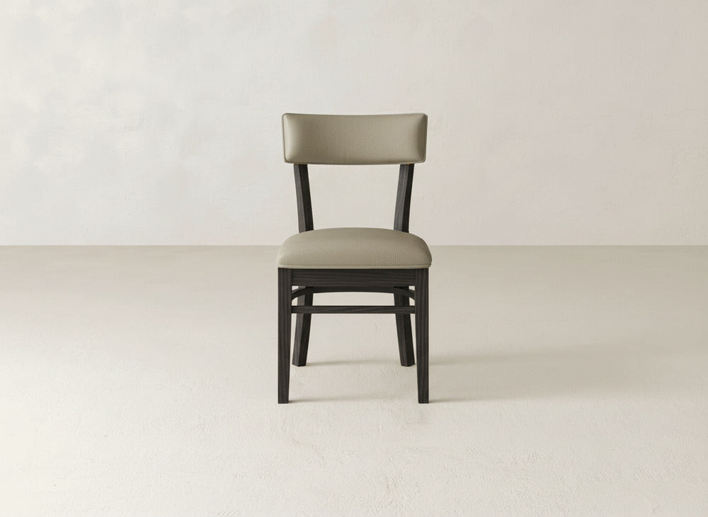 The Everly Side Dining Chair by James+James, featuring a cushioned seat and backrest in light beige upholstery, is centered against a plain, light-toned wall and floor.