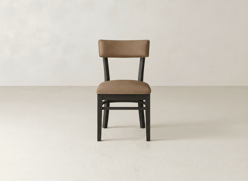 The Everly Side Dining Chair by James+James features brown upholstery and a dark wood frame, inspired by mid-century design, shown facing forward on a light-colored floor against an off-white wall.