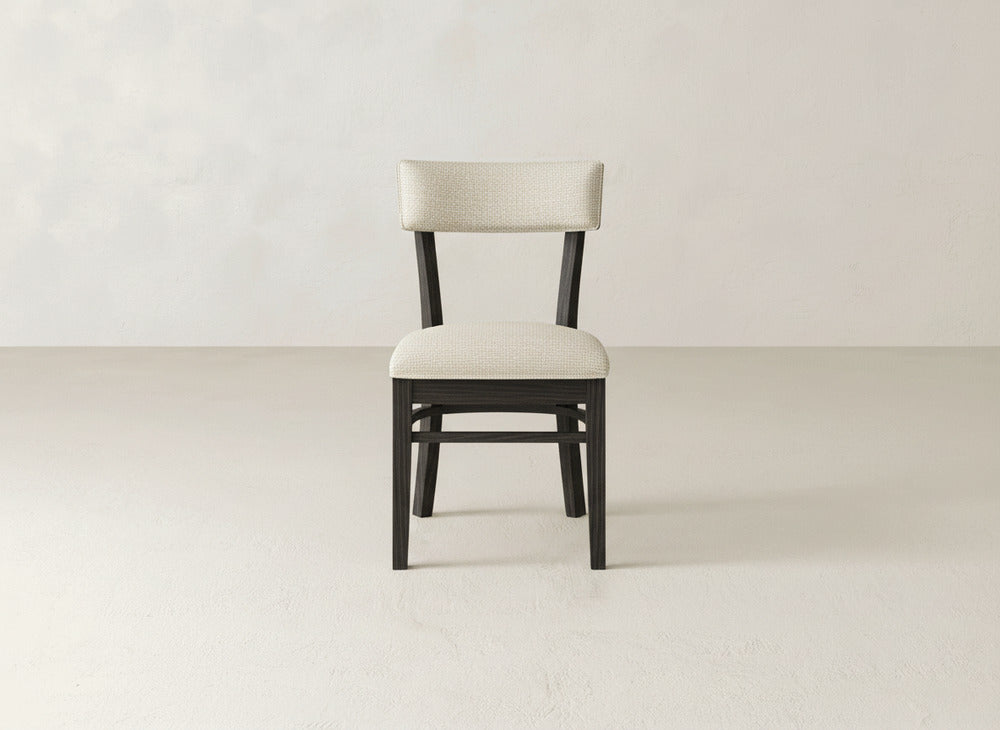 The Everly Side Dining Chair by James+James, featuring a dark frame and light upholstered seat and backrest, is centered against a plain, light-toned wall and floor.